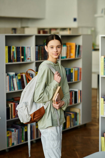 Een meisje met een rugzak staat in een bibliotheek, verdiept in haar omgeving terwijl ze de boekenplanken verkent. - Foto, afbeelding