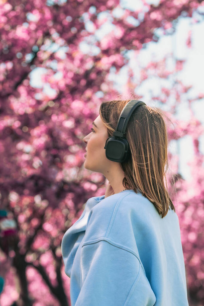 stijlvolle vrouw meloman luisteren muziek kopiëren ruimte bloeiende sakura op de achtergrond - Foto, afbeelding