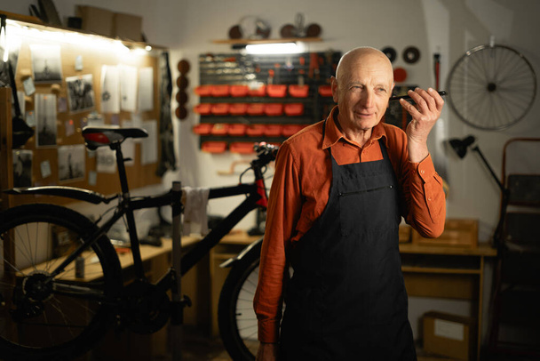 Old man male bicycle mechanic listening to voice message while standing in garage or bicycle repair shop. Copy space - Photo, Image