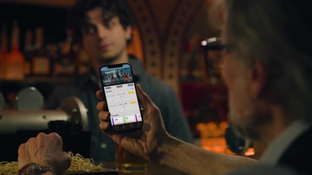 Mature sports fan bets on football match online using mobile phone, talks with bartender and friend. Man checks game score and bookmaker ratings in mobile app sitting at bar counter in pub. Gambling - Footage, Video
