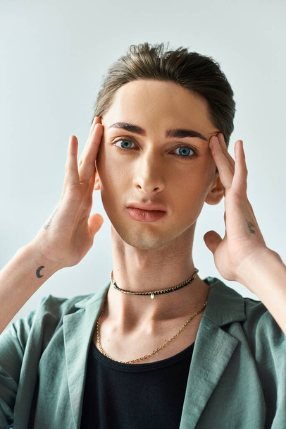 Ένας νεαρός queer άνθρωπος στέκεται με τα χέρια στο κεφάλι, βαθιά σε περισυλλογή, σε ένα στούντιο με γκρι φόντο. - Φωτογραφία, εικόνα