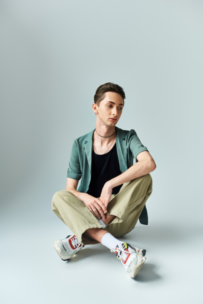 Un giovane omosessuale siede sul pavimento, indossa una giacca verde e scarpe da ginnastica, trasudando fiducia e orgoglio in un ambiente da studio.. - Foto, immagini