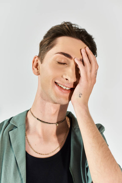 Ein junger queerer Mensch lächelt hell, die Hände auf dem Gesicht, verströmt Stolz und Zuversicht auf grauem Studiohintergrund. - Foto, Bild