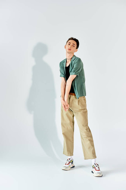 A young queer person strikes a stylish pose in a tan shirt and khaki pants, exuding confidence in front of a white background. - Photo, Image