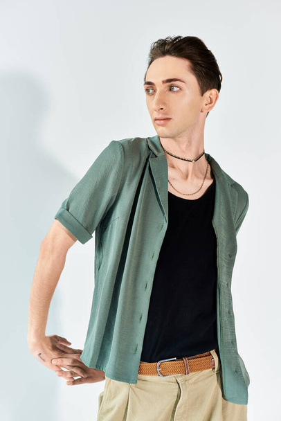 Une jeune queer pose en toute confiance dans un studio portant une chemise verte et un pantalon bronzé sur fond gris. - Photo, image