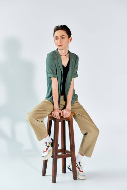 Un jeune queer s'assoit sur un tabouret dans un studio, posant sur un fond gris avec un sentiment de fierté et d'authenticité. - Photo, image