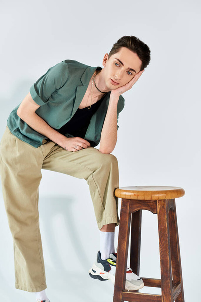 queer person, part of the LGBTQ+ community, sits proudly on a stool in khaki pants and a green shirt against a grey background. - Photo, Image