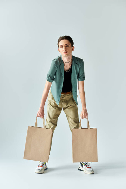 Un joven queer sosteniendo dos bolsas de la compra sobre un fondo gris, expresando alegría y orgullo en sus compras. - Foto, imagen