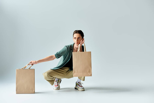 Un joven queer se agacha, exudando orgullo, sosteniendo bolsas de compras en un estudio sobre un fondo gris. - Foto, imagen