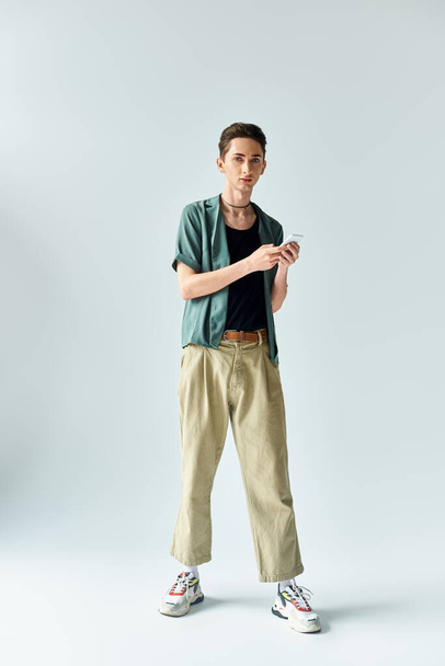 Молодой человек в рубашке и брюках цвета хаки, держащий мобильный телефон, излучающий гордость и уверенность в студийной обстановке. - Фото, изображение