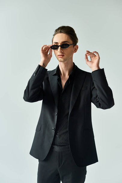Een jonge homo slaat een zelfverzekerde pose in een zwart pak en een zonnebril tegen een grijze achtergrond. - Foto, afbeelding