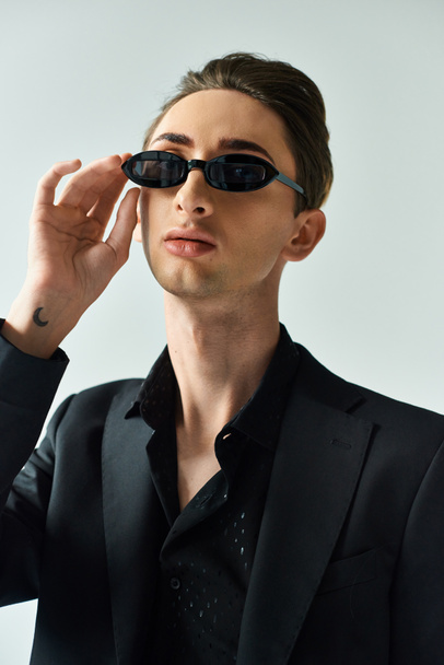 Un jeune queer prend une pose confiante en costume pointu, portant des lunettes de soleil sur un fond gris. - Photo, image