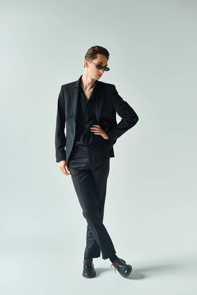 Uma jovem queer pessoa em um terno preto elegante marcando uma pose confiante em um estúdio contra um fundo cinza. - Foto, Imagem
