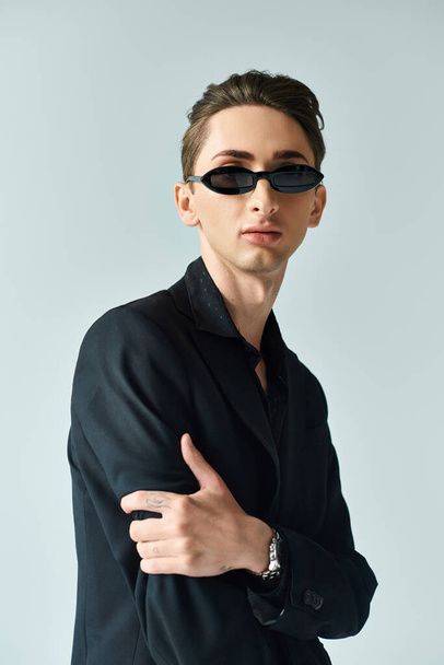 Uma jovem queer pessoa em um terno elegante e óculos de sol golpeia uma pose confiante contra um pano de fundo cinza. - Foto, Imagem