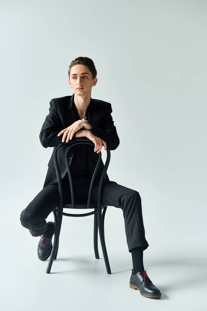 Uma jovem queer senta-se confiantemente em um terno em uma cadeira contra um fundo cinza, exalando orgulho e empoderamento. - Foto, Imagem