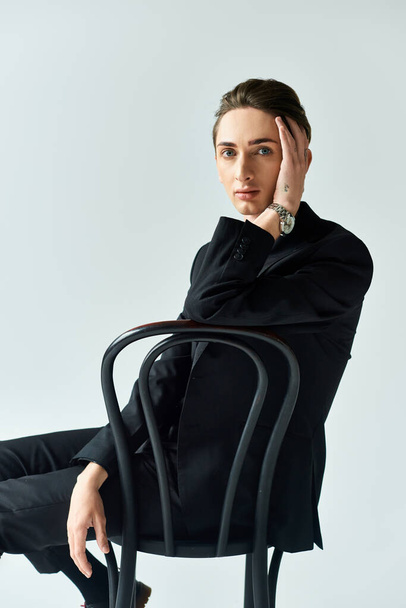 Ein junger queerer Mensch, der Zuversicht ausstrahlt, sitzt auf einem Stuhl im schicken schwarzen Anzug vor grauem Studiohintergrund.. - Foto, Bild