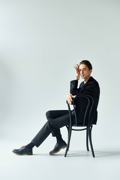 Um jovem elegante de terno se senta em uma cadeira, exalando confiança e contemplação em um estúdio com um fundo cinza. - Foto, Imagem