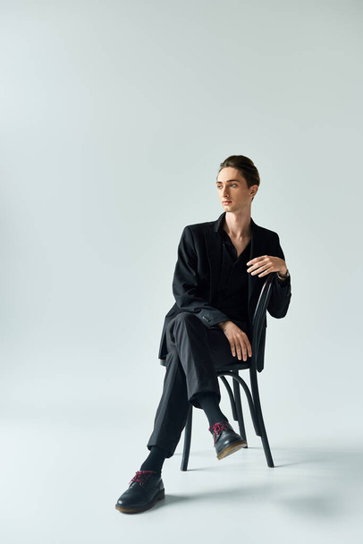 Młody mężczyzna w czarnym garniturze emanuje pewnością siebie siedząc na krześle w studio na szarym tle. - Zdjęcie, obraz