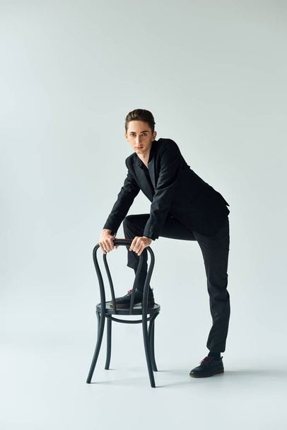 Молодий квір-чоловік у стильному костюмі впевнено спирається на стілець у студійній обстановці з сірим фоном. - Фото, зображення