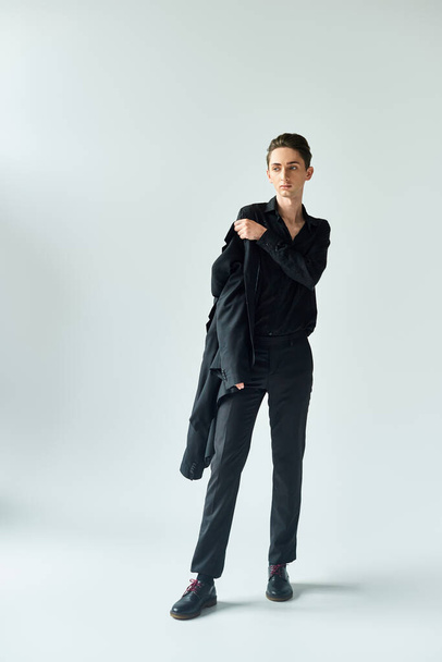 Ein junger queerer Mensch, der Stolz ausstrahlt, posiert in einem Studio mit schwarzem Mantel und Hose vor grauem Hintergrund. - Foto, Bild