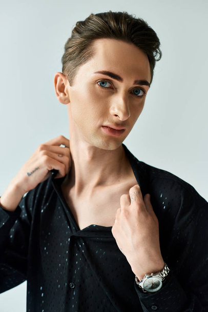 Um jovem com uma camisa preta ajusta seu colarinho, exalando confiança e estilo, em um estúdio contra um fundo cinza. - Foto, Imagem