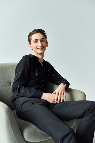 A young queer man sits confidently in a chair, wearing a black shirt, showcasing pride and strength in a studio setting on a grey background. - Photo, Image