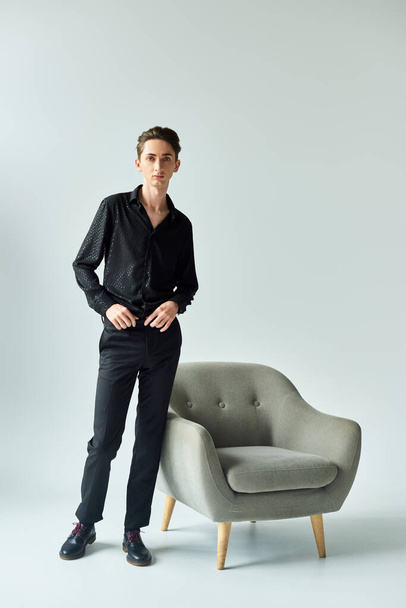 Una giovane persona queer con una camicia nera che colpisce una posa accanto a un'elegante sedia grigia in un ambiente da studio. - Foto, immagini