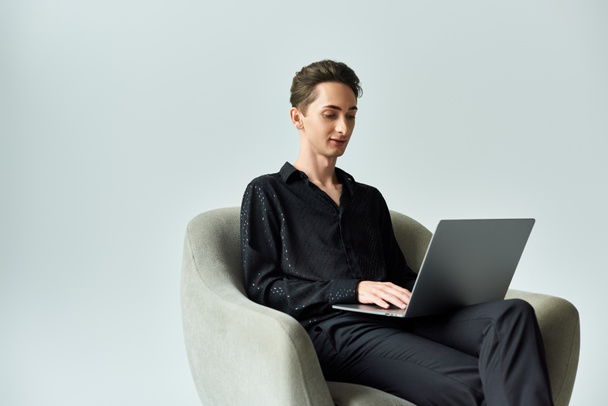 Une jeune personne queer, exprimant fièrement son identité, assise sur une chaise à l'aide d'un ordinateur portable dans un studio avec un fond gris. - Photo, image