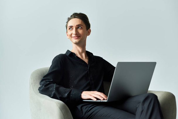 Een jonge homo op een grijze achtergrond zit in een stoel met een laptop en straalt vertrouwen en trots uit in hun digitale aanwezigheid.. - Foto, afbeelding
