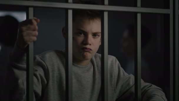Valkoihoinen teini, jolla on kasvotatuoinnit, seisoo sellissä vankilassa, pitää metallibaareja ja katsoo kameraa. Monipuoliset nuoret vangit puhuvat taustalla. Nuorisovankila tai rangaistuslaitos - Materiaali, video
