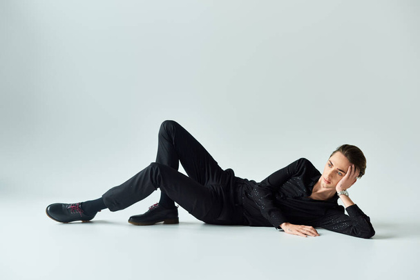 Ένας νεαρός ομοφυλόφιλος ποζάρει κομψά με μαύρο κοστούμι σε ένα γκρίζο πάτωμα στούντιο, αποπνέοντας αυτοπεποίθηση και υπερηφάνεια.. - Φωτογραφία, εικόνα