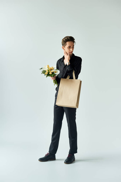 Un jeune queer frappe une pose tenant un sac à provisions et un bouquet de fleurs sur un fond de studio gris. - Photo, image