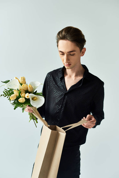 Een jonge homo houdt trots een papieren zak vol bloemen in een studio tegen een grijze achtergrond. - Foto, afbeelding