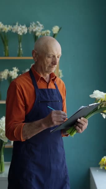 Concepto de venta, negocio y floristería. Viejo hombre florista haciendo notas en el portapapeles, haciendo lista de verificación de inventario trabajando en floristería. Vista vertical - Imágenes, Vídeo
