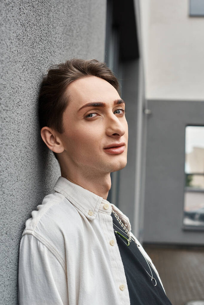 Un joven con una identidad queer se apoya contemplativamente contra una pared, exudando estilo y confianza. - Foto, Imagen