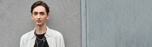 Un giovane omosessuale in abiti eleganti si erge audacemente di fronte a un muro grigio, irradiando orgoglio e sfida.. - Foto, immagini
