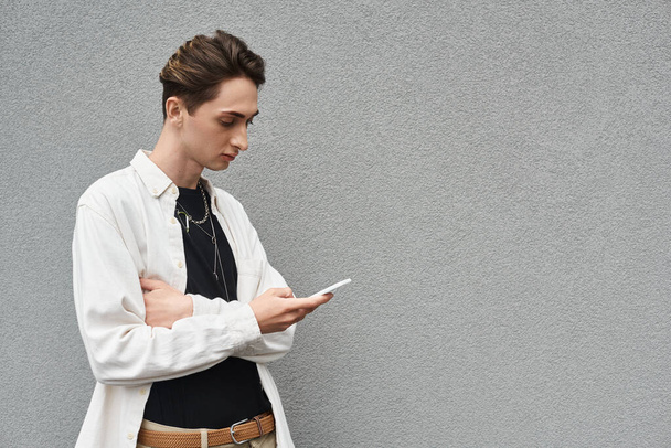 Elegante giovane queer in abbigliamento casual appoggiato a un muro, in possesso di un telefono cellulare. - Foto, immagini