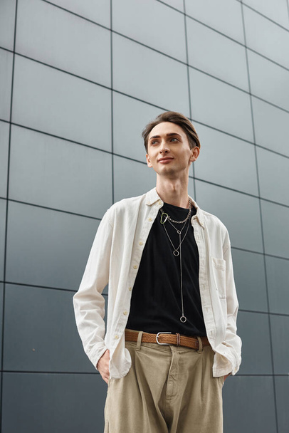Un jeune queer vêtu d'une chemise blanche et d'un pantalon bronzé pose en toute confiance devant un immeuble. - Photo, image