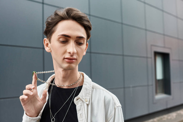 Стильный молодой человек держит ожерелье перед величественным зданием, демонстрируя свое уникальное чувство моды и гордости. - Фото, изображение
