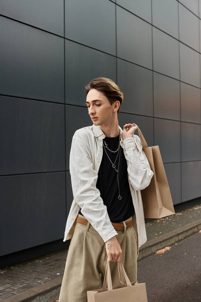 Un joven LGBTQ con un atuendo elegante llevando bolsas de compras frente a un edificio moderno. - Foto, Imagen