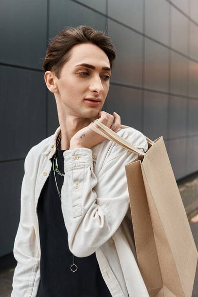Ένα κομψό νεαρό άτομο με μια τσάντα ψώνια στέκεται μπροστά από ένα σύγχρονο κτίριο. - Φωτογραφία, εικόνα