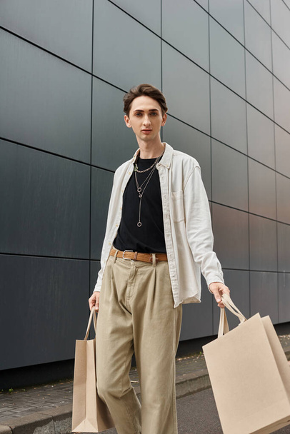 Ένα νεαρό queer άτομο με κομψή ενδυμασία, κρατώντας τσάντες για ψώνια, στέκεται μπροστά από ένα σύγχρονο κτίριο. - Φωτογραφία, εικόνα