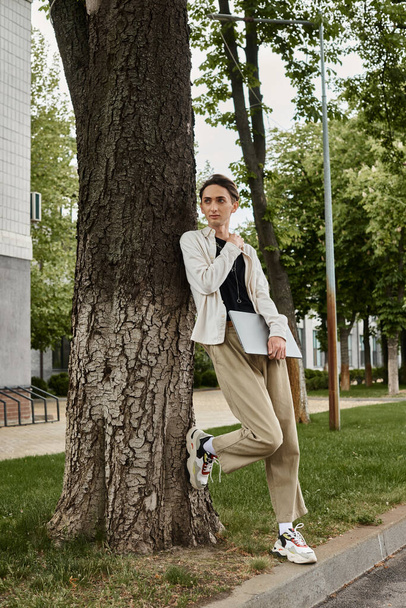 Ein junger queerer Mensch in stilvoller Kleidung lehnt nachdenklich an einem Baum und verkörpert Stolz und Besinnlichkeit. - Foto, Bild