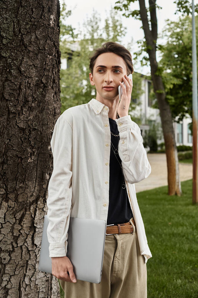 Egy fiatal, elegánsan öltözött buzi, aki egy fa mellett áll, és a telefonján cseveg egy alkalmi szabadtéri környezetben.. - Fotó, kép