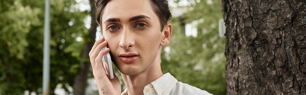 Nuori homo yksilö muodikkaissa vaatteissa keskustelee matkapuhelimessa puun edessä, johon sisältyy ylpeys ja kuuluminen.. - Valokuva, kuva