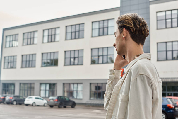 Ένα νεαρό ομοφυλόφιλο άτομο με μοντέρνα ενδυμασία που μιλάει στο κινητό του μπροστά από ένα αστικό κτίριο, επιδεικνύοντας την περηφάνια των ΛΟΑΤΚΙ. - Φωτογραφία, εικόνα