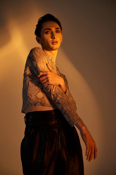 Une jeune queer en pantalon en cuir noir frappe une pose confiante devant une lumière lumineuse. - Photo, image