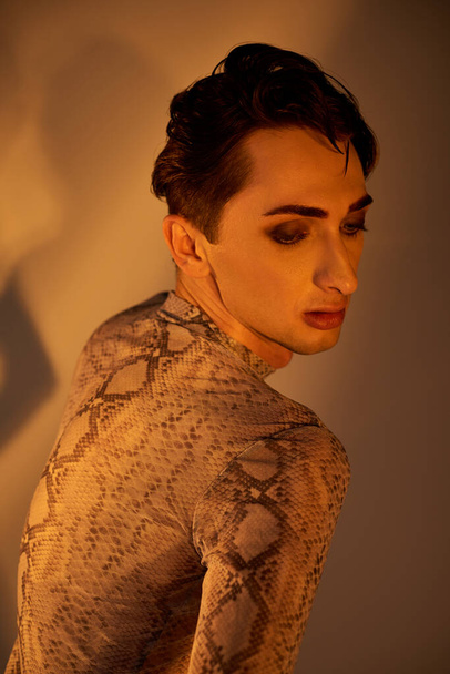 Ένας νεαρός queer άνθρωπος ποζάρει μπροστά από έναν καθρέφτη φορώντας ένα φόρεμα δέρμα πύθωνα, αποπνέοντας αυτοπεποίθηση και στυλ. - Φωτογραφία, εικόνα