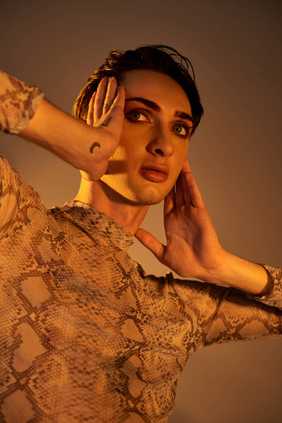 Una joven queer en un vestido de piel de serpiente posa con las manos en las orejas en un elegante conjunto inspirado en el orgullo. - Foto, imagen