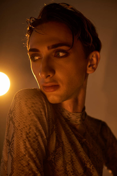 Een jonge homo in stijlvolle kledij staat vol vertrouwen voor een stralend licht in een schemerige kamer. - Foto, afbeelding